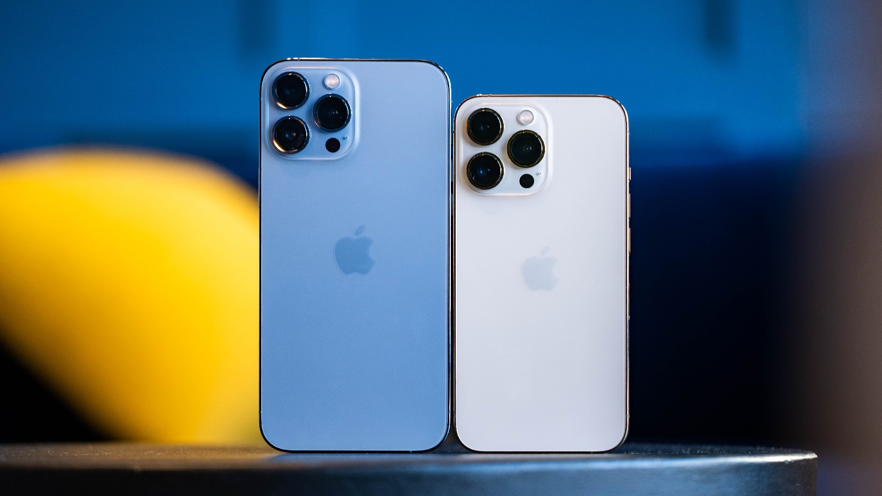 Een blauwe iPhone 13 Pro Mac en een gouden iPhone 13 Pro naast elkaar op een tafel met hun achterste camera's naar ons gericht.
